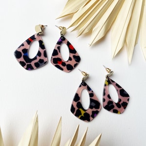 May include: A pair of pink, black, and multi-colored leopard print earrings with gold accents. The earrings are shaped like a teardrop with a large oval cutout in the center.