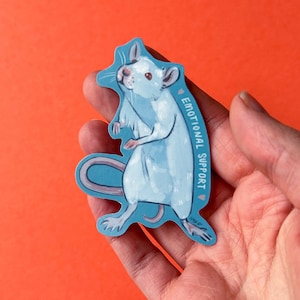 Könnte beinhalten: Ein blauer und weißer Aufkleber mit einer Cartoon-Ratte und dem Text "Emotional Support" sowie einem kleinen rosa Herz.