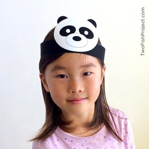 May include: A child wearing a black headband with a white panda face cutout. The panda face has black eyes and a black nose.