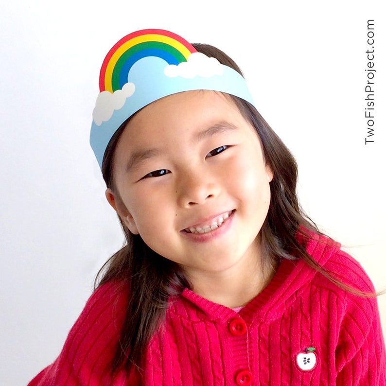 May include: A child wearing a red sweater with an apple design is wearing a paper rainbow headband with clouds. The headband is blue and white with a rainbow of red, orange, yellow, green, blue, indigo, and violet.