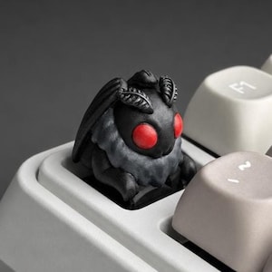 May include: A custom artisan keycap shaped like a moth with a matte black body, textured wings, and two prominent, smooth, bright red circular eyes, installed on a mechanical keyboard next to several light gray keys with white lettering.