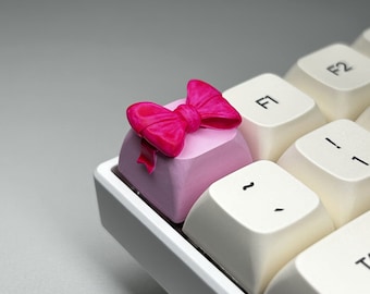 May include: A pink keyboard keycap with a pink bow on top. The keycap is on a white keyboard.