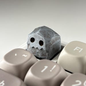 May include: A custom mechanical keyboard keycap shaped like a textured gray rock with two circular eye cutouts and a jagged mouth opening. The keycap is positioned on a keyboard among light beige keys, including one labeled with F1 and 1!.