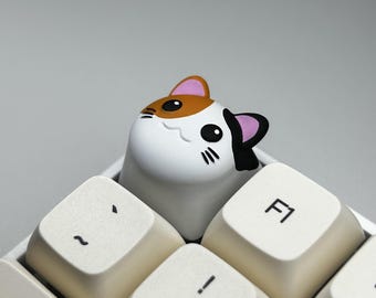 May include: A custom cat-shaped keycap in white, brown, and black, placed on a computer keyboard. The keycap has a detailed design of a cat's face. The keyboard keys are off-white with black lettering.