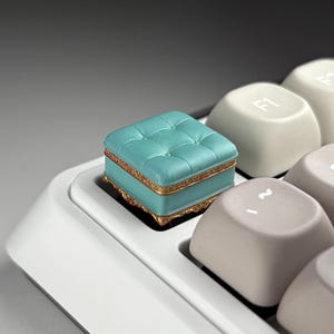 May include: A teal and gold decorative keyboard keycap shaped like a miniature ottoman. The keycap has a tufted design and sits on a white keyboard. The keycap has gold trim.