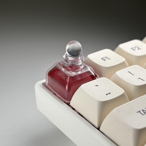 May include: A small, clear glass bottle filled with a deep red liquid, topped with a spherical stopper. The bottle sits on a white keyboard, positioned between the keys. The keys are off-white with black lettering.