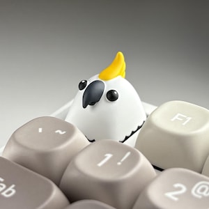 May include: A white parrot figurine with a yellow crest sits among computer keyboard keys. The keys are various shades of gray and beige, with white lettering. The parrot has black eyes and a black beak.