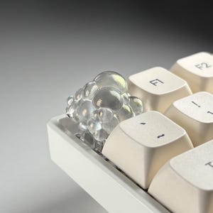 May include: A clear, bubble-shaped artisan keycap sits on a white mechanical keyboard. The keycap has an iridescent sheen, reflecting various colors. The surrounding keys are off-white with black lettering, including the F1 and F2 keys.