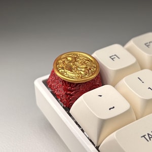 May include: A custom keyboard keycap with a gold coin design atop a red, ornate base. The keycap is installed on a white keyboard, with other keys visible in the background. The coin features a detailed relief design.
