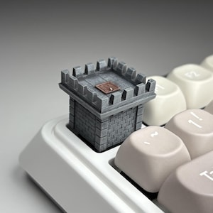 May include: A gray, miniature castle tower keycap, with a brick pattern and a small door on top, sits atop a white keyboard. The keycap is a detailed, 3D-printed accessory, adding a unique aesthetic to a mechanical keyboard.