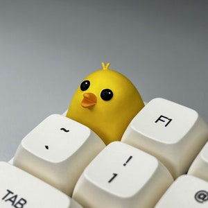 Peut inclure: Une figurine de poussin jaune est posée sur un clavier blanc avec des touches étiquetées "F1", "!" et "TAB".