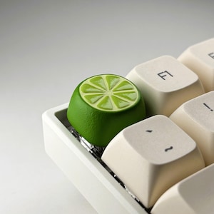 Pode incluir: Uma tecla mecânica personalizada em forma de fatia de lima, apresentando um exterior verde vibrante e texturizado e um padrão detalhado de segmentos de citrinos em verde claro no topo, ao lado de teclas em tom creme com texto em preto.