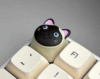 May include: A custom keyboard keycap featuring a Siamese cat design. The keycap is cream-colored with a black and brown cat face, blue eyes, and pink ears. The keycap is positioned among other cream-colored keys with black lettering.