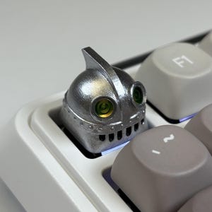 May include: A silver robot-themed keycap installed on a white and gray mechanical keyboard. The keycap has a detailed design with green eye accents and a mouth-like vent. The keyboard keys are visible in the foreground.