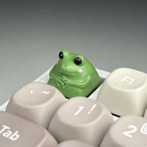 Puede incluir: Una tecla con forma de rana verde, con ojos negros y mejillas rosas, entre teclas beige de teclado. Esta peculiar tecla añade un toque divertido a un teclado mecánico.