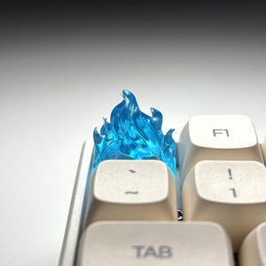 May include: A translucent blue flame-shaped keycap sits atop a white keyboard. The keycap is positioned between the Tab, F1, and number 1 keys. The flame design is detailed and appears to be made of a clear, glossy material.