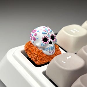 May include: A white skull-shaped keycap with pink and blue floral designs and black eye sockets. The keycap is surrounded by an orange floral garland and sits on a white keyboard.