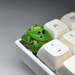 May include: A green and yellow keycap shaped like a cartoon monster with black eyes. The keycap is sitting on a white keyboard.