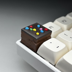 May include: A chocolate cake keycap with colourful sprinkles on top, sitting on a white keyboard with F1 and F2 keys visible.