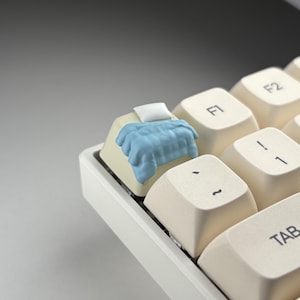 Könnte beinhalten: Nahaufnahme einer Computertastatur mit einer individuellen Tastenkappe. Die Tastenkappe ist beige und zeigt ein Miniaturbett mit einer blauen Decke und einem weißen Kissen. Weitere Tasten sind sichtbar, darunter F1, F2 und Tab.