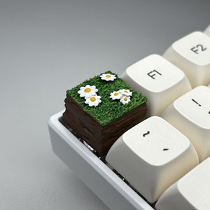 May include: A small, brown, wooden block with green grass and white flowers on top. The block is sitting on a white keyboard.