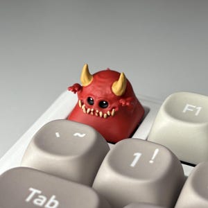 Puede incluir: Una tecla roja con forma de diablo con cuernos amarillos y una sonrisa dentuda se encuentra en un teclado blanco. La tecla está entre las teclas Tab, 1! y F1. Las teclas del teclado son de color gris claro.
