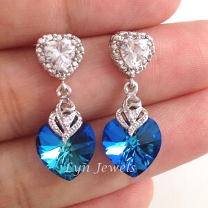May include: A pair of silver heart-shaped earrings with a dangling blue crystal heart. The top heart is encrusted with clear rhinestones.