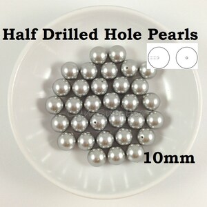 Puede incluir: Un bol de perlas de plata de 10 mm con un agujero perforado a la mitad. Las perlas son redondas y tienen un acabado liso y brillante. La imagen muestra un primer plano de las perlas, con el texto "Half Drilled Hole Pearls" y "10mm" visible.