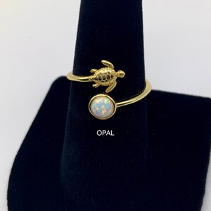 May include: Gold ring with a small turtle charm and a round opal gemstone. The ring is on a black ring display stand. "OPAL" is written below the ring.