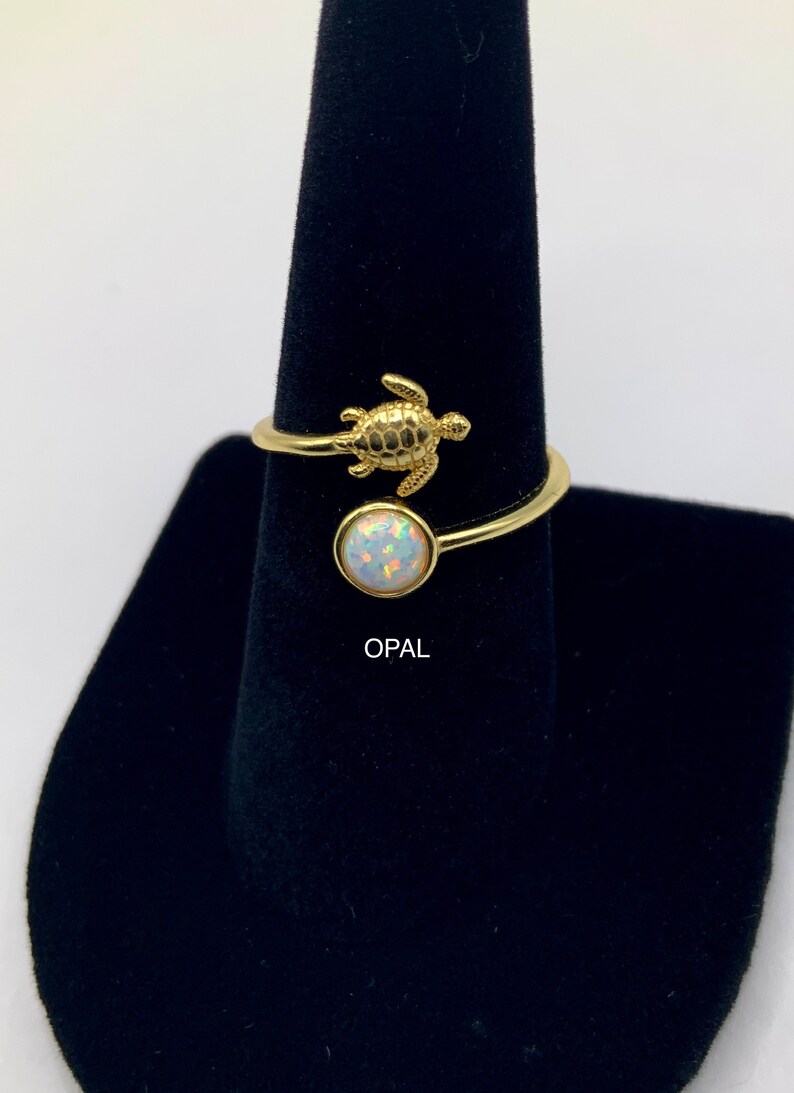 May include: Gold ring with a small turtle charm and a round opal gemstone. The ring is on a black ring display stand. "OPAL" is written below the ring.