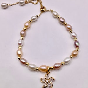 Könnte beinhalten: Ein verstellbares Armband aus Gold mit einem Blumenanhänger, der mit klaren Zirkonia-Steinen besetzt ist. Das Armband besteht aus einer Reihe von weißen, rosa und pfirsichfarbenen Perlen.