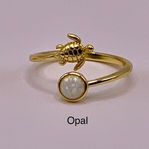 May include: Three gold rings with a turtle charm and a gemstone. The rings are available with a blue opal, white opal, and a light blue larimar stone.