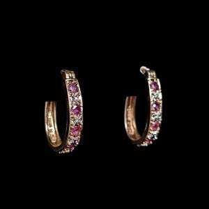 May include: A pair of gold hoop earrings featuring a row of round-cut pink gemstones alternating with small clear white stones. The earrings have a hinged closure mechanism and a polished metallic finish, set against a solid black background.