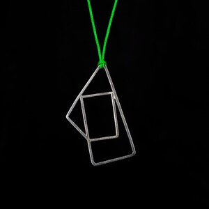 Puede incluir: Un collar con un colgante geométrico de plata en un cordón verde. El colgante está hecho de dos formas superpuestas, un triángulo y un rectángulo.