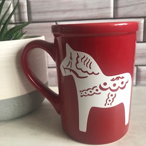 May include: Red ceramic mug with a white Dala horse design. The horse is a traditional Swedish folk art motif.