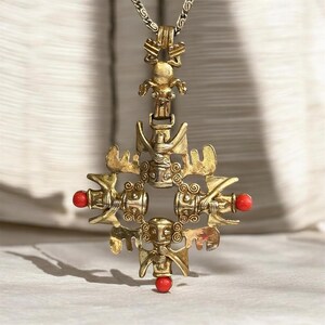 May include: A gold-toned metal pendant necklace featuring a central cross-shaped design composed of four stylized figures with wings. The pendant is accented with three small, round, bright red beads at the left, right, and bottom points of the cross.