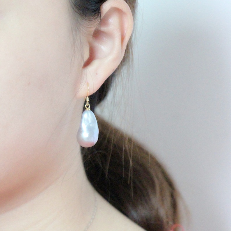 May include: A pair of silver drop earrings featuring a large, irregular-shaped pearl. The pearl has a slight pink hue and is suspended from a gold-toned hook.