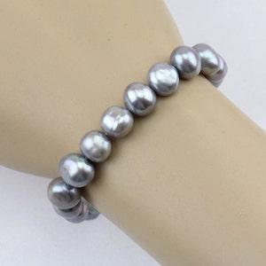 May include: A bracelet featuring a strand of large, gray pearls. The pearls are round and have a lustrous sheen. The bracelet is worn on a wrist, showcasing the jewelry's elegant design. The pearls are approximately 0.5 inches in diameter.