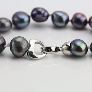 May include: A close-up of a necklace featuring dark gray baroque pearls. The pearls are strung together with a silver-colored heart-shaped clasp. The pearls have iridescent hues of green, purple, and blue.