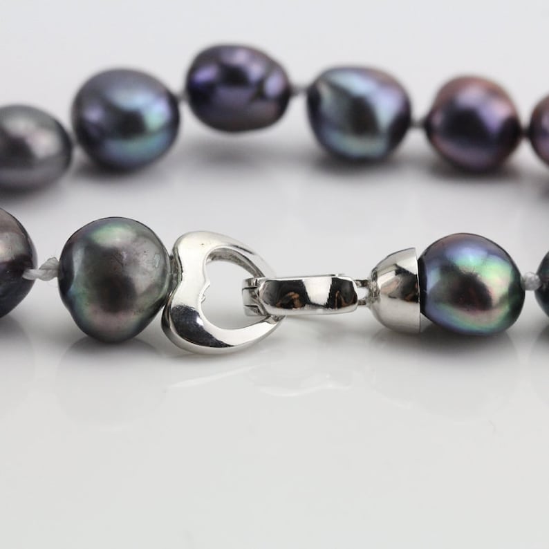 May include: A close-up of a necklace featuring dark gray baroque pearls. The pearls are strung together with a silver-colored heart-shaped clasp. The pearls have iridescent hues of green, purple, and blue.