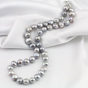 May include: A necklace of gray freshwater pearls with a silver clasp, resting on a white satin fabric. The pearls are irregularly shaped and have a lustrous sheen, creating a classic and elegant look.
