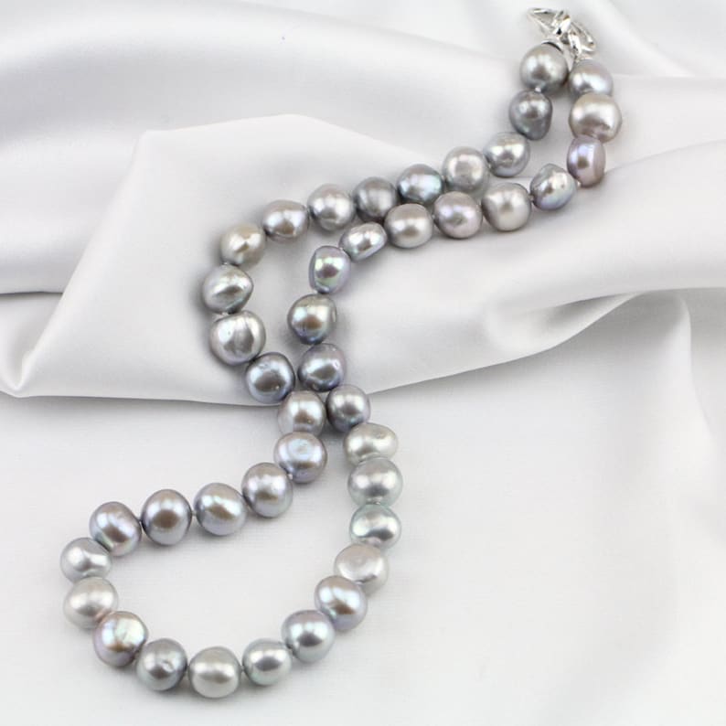 May include: A necklace of gray freshwater pearls with a silver clasp, resting on a white satin fabric. The pearls are irregularly shaped and have a lustrous sheen, creating a classic and elegant look.
