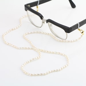 May include: A white pearl eyeglass chain with gold clasps. The chain is looped over a pair of black framed glasses.