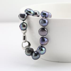 May include: A dark gray pearl bracelet with a silver clasp. The pearls are irregularly shaped and have iridescent hues of green and purple. The bracelet is displayed against a white background.