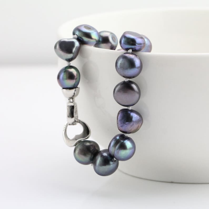 May include: A dark gray pearl bracelet with a silver clasp. The pearls are irregularly shaped and have iridescent hues of green and purple. The bracelet is displayed against a white background.