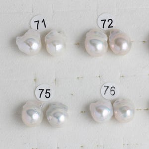 May include: Four pairs of white, irregular-shaped baroque pearls arranged in two rows. Each pair is labeled with a small white circular sticker featuring black numbers 71, 72, 75, and 76, indicating distinct sets of lustrous, organic-shaped pearls.