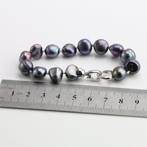 May include: A bracelet featuring dark gray baroque pearls with iridescent hues. The pearls are strung together and secured with a silver-colored clasp. A ruler is placed next to the bracelet for measurement.