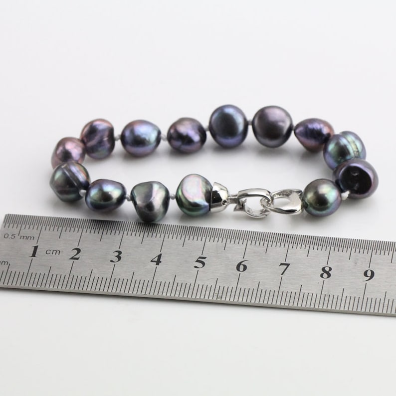 May include: A bracelet featuring dark gray baroque pearls with iridescent hues. The pearls are strung together and secured with a silver-colored clasp. A ruler is placed next to the bracelet for measurement.