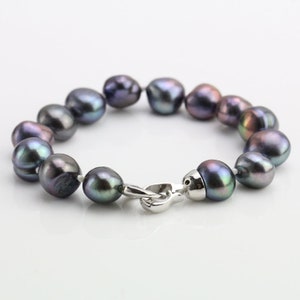 May include: A bracelet featuring iridescent dark gray pearls strung together. The pearls have a smooth, rounded shape and are connected with a silver-colored clasp. The bracelet is displayed on a white surface.