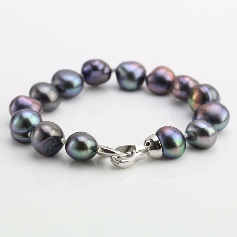 May include: A bracelet featuring iridescent dark gray pearls strung together. The pearls have a smooth, rounded shape and are connected with a silver-colored clasp. The bracelet is displayed on a white surface.
